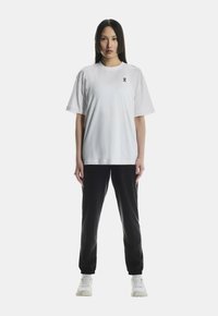 Kvinde står frontvendt iført en hvid oversize t-shirt, sorte joggers og hvide sneakers foran en ensfarvet baggrund.