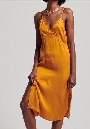 Frau trägt ein senfgelbes Satin-Nachthemd mit Spaghettiträgern, V-Ausschnitt, Seitenschlitz und mehreren silbernen Halsketten.