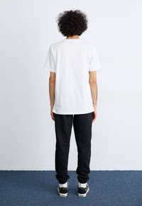 Weißes Baumwoll-T-Shirt, runder Ausschnitt, kurze Ärmel, kombiniert mit schwarzen Jogginghosen, elastischen Bündchen und weißen Akzent-Sneakern. Rückansicht.