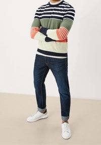 Mann in einem bunten gestreiften Langarmpullover, dunklen hochgekrempelten Jeans und weißen Turnschuhen, der mit verschränkten Armen steht.