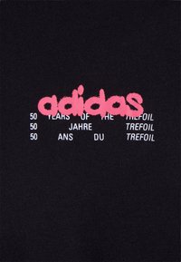 Svart tyg med rosa "adidas"-text överlagrat på vit text som läser "50 år av treklövern" på engelska, tyska och franska.