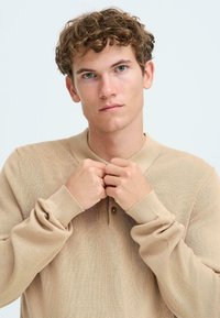 Beige strikkegenser med lange ermer, kragehals og knappedetalj. Myk tekstur med lett utseende.