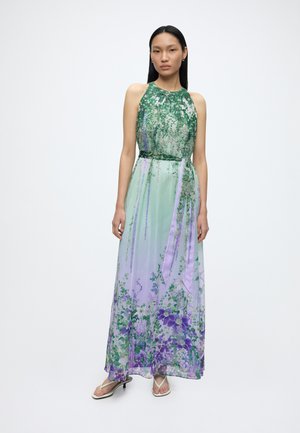 Woman wearing a sleeveless green and lavender floral maxi dress with a high neckline and thin heeled sandals, standing against a white background.