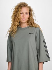 Junge Person mit langen lockigen blonden Haaren, die ein übergroßes graues Hummel-Sportshirt mit schwarzen Chevron-Streifen auf dem Ärmel trägt.