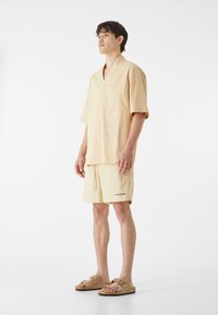 Svetlo béžová krátka tričko s výstrihom do V, spárované s rovnakými šortkami. Text na šortkách znie "SKIN SENSE." Model má na sebe hnedé sandále.