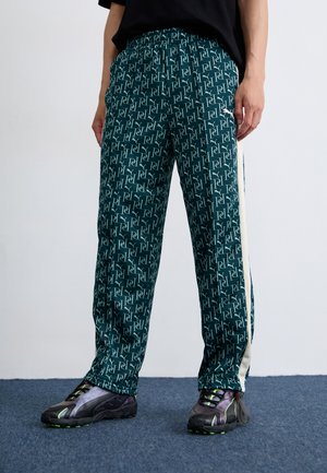 A person wearing patterned green pants and black shoes stands on a blue carpeted floor against a plain white wall.