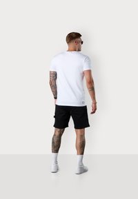 Weißes T-Shirt mit kurzen Ärmeln und Rundhalsausschnitt, kombiniert mit schwarzen Shorts und weißen Sneakers. Tätowierungsdetails sind an Armen und Beinen sichtbar.