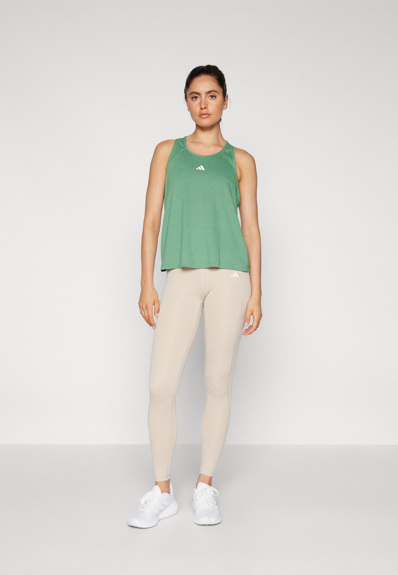 Top verde sin mangas con cuello redondeado y logo blanco, combinado con leggins beige y zapatillas blancas. Tejido suave, diseño ajustado.