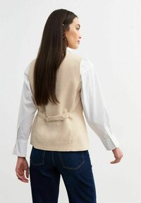 Chaleco de lino beige con un respaldo entallado, que presenta un cinturón estrecho en la cintura. Combinado con una camisa blanca de manga larga y jeans de mezclilla azul oscuro.