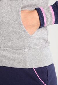 Main dans la poche d'un sweat-shirt gris avec un poignet rayé ; pantalon bleu marine avec un passepoil rose visible à la taille.
