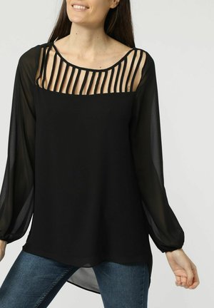 Mujer con una blusa negra transparente de mangas largas con detalles de tiras verticales en el escote y vaqueros azules, sonriendo levemente.