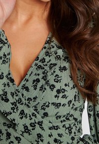Green, textured fabric top with a deep V-neck and black floral patterns. Ruffled neckline detail and long sleeves visible.