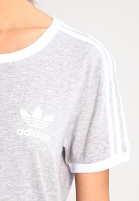 Šedé bavlněné tričko s bílými akcenty, kulatý výstřih, krátké rukávy a tři bílé pruhy na ramenou. Obsahuje bílé logo Adidas.