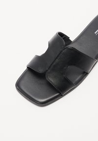 Sandale noire à semelle plate avec un bout carré, présentant un dessus en cuir lisse et deux larges sangles pour un maintien sécurisé. Semelle intérieure texturée pour le confort.