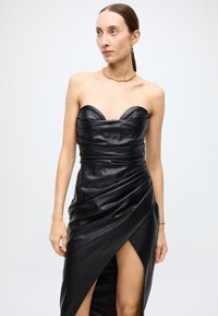 Black leather dress with a strapless design, draped texture, and a high slit. Features a curved neckline and smooth, shiny surface.