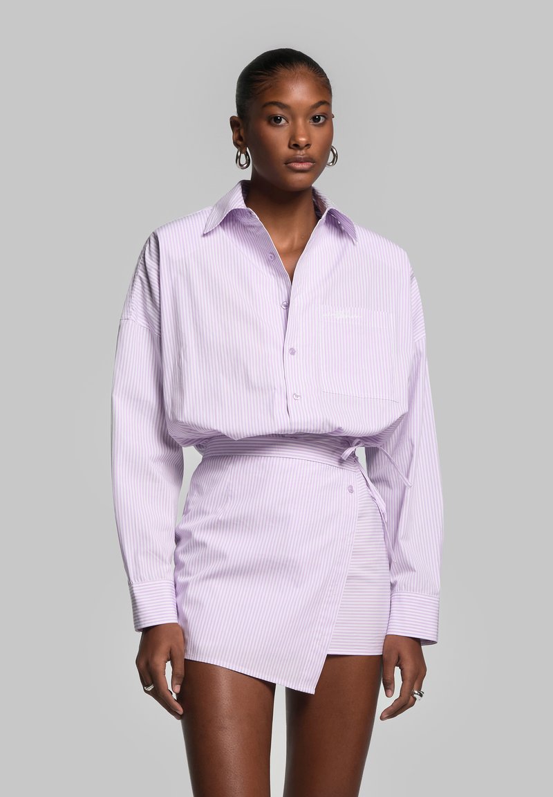 Chemise boutonnée rayée lilas avec une poche, associée à une jupe portefeuille assortie. Texture medium, coupe décontractée, manches retroussées et détails de boutons.