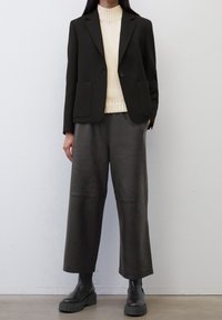 Blazer noir avec deux poches avant, col roulé texturé crème, et pantalon large en cuir noir. De grosses bottes noires complètent la tenue.
