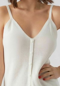 Top in maglia bianca con spalline sottili, scollatura a V e bottoni frontali. Il tessuto presenta un motivo testurizzato ed è leggero.