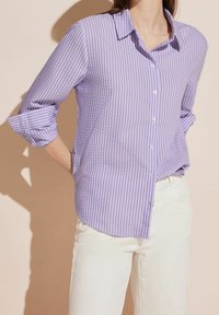 Ljuslila randig skjorta med krage, uppvikta ärmar och vita knappar, tillsammans med krämfärgade byxor.