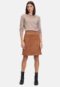 Gonna A-line in suede marrone con due bottoni laterali. Abbinata a un maglione beige a girocollo e stivaletti neri. Tessuto liscio e forma aderente.