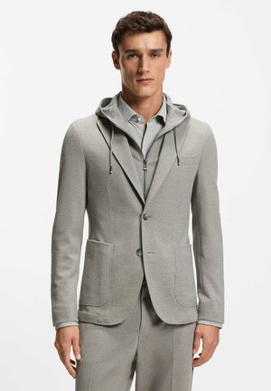 Hombre con chaqueta de traje gris claro a medida con bolsillos delanteros, sobre una sudadera con capucha con cremallera y camisa con cuello, posando sobre un fondo blanco.