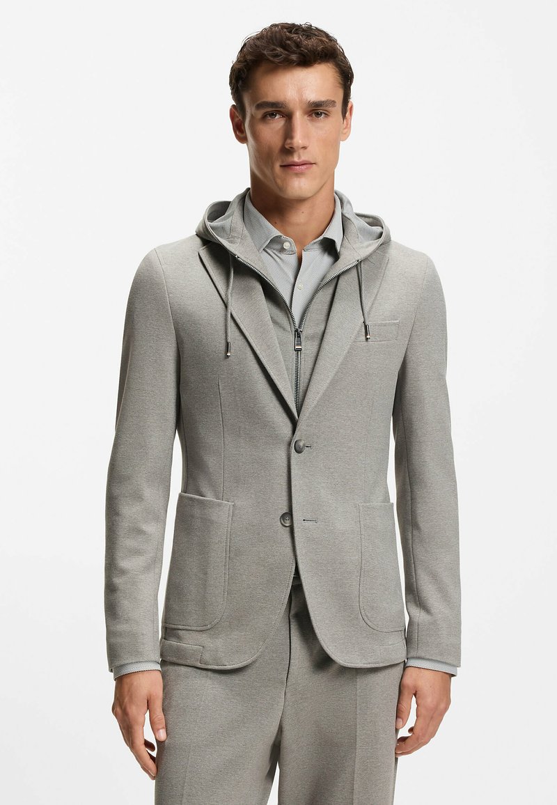 Mężczyzna w jasnoszarej, dopasowanej marynarce z przednimi kieszeniami, noszący pod nią bluzę na zamek i koszulę z kołnierzykiem, stoi na tle białego tła.