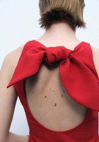 Il vestito rosso presenta un design con schiena scoperta e un grande fiocco dettaglio al collo, realizzato in tessuto morbido, che mette in evidenza la texture e le macchie della pelle.
