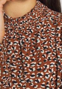 Brun blus i tyg med smockad halsringning, prydd med ett spritt djurmönster i svart, vitt och teal. Strukturerad yta.