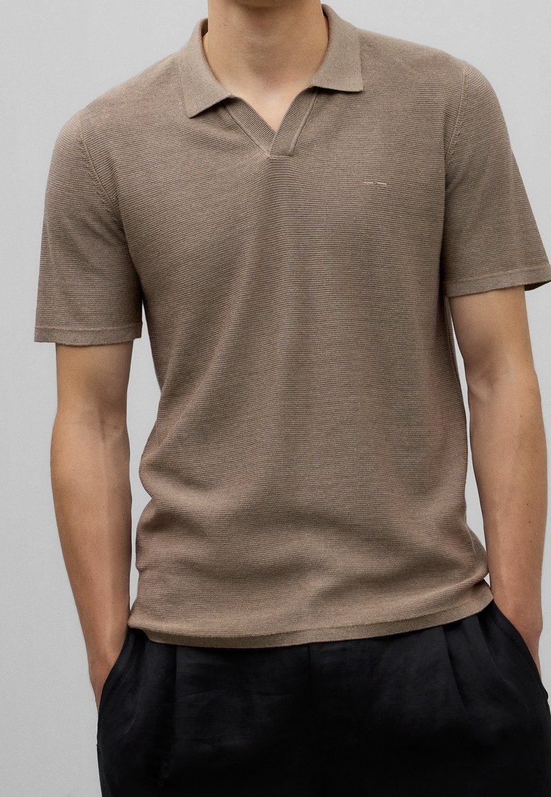 Homme portant un polo en maille marron à manches courtes avec col et un pantalon noir, debout les mains dans les poches devant un fond uni.