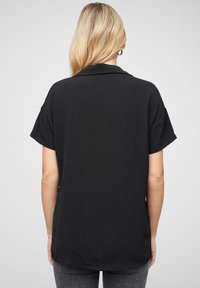 Camicia nera a maniche corte con colletto, realizzata in un materiale testurizzato. Presenta una vestibilità comoda e un'orlo posteriore leggermente più lungo.