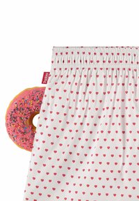 Hvit stoff med små røde hjerter, elastisk midjebånd, og en rosa donut med fargerike strøssel delvis synlig på siden.