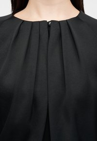 Gros plan sur l'encolure d'un vêtement noir avec des plis verticaux et un petit bouton noir au centre devant.