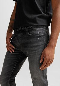 Jeans de denim negros para hombre con corte ajustado, que presentan una textura desvanecida, un mínimo desgaste, diseño estándar de cinco bolsillos y cierre de botón.