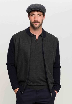 Dark gray zip-up cardigan with navy blue ribbed sleeves, paired with a dark shirt. The model wears a gray flat cap, posing against a white background.