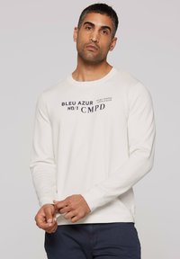 Long-sleeve, off-white t-shirt made of cotton blend, featuring navy text and a crew neck. Hands adjusting the hem.