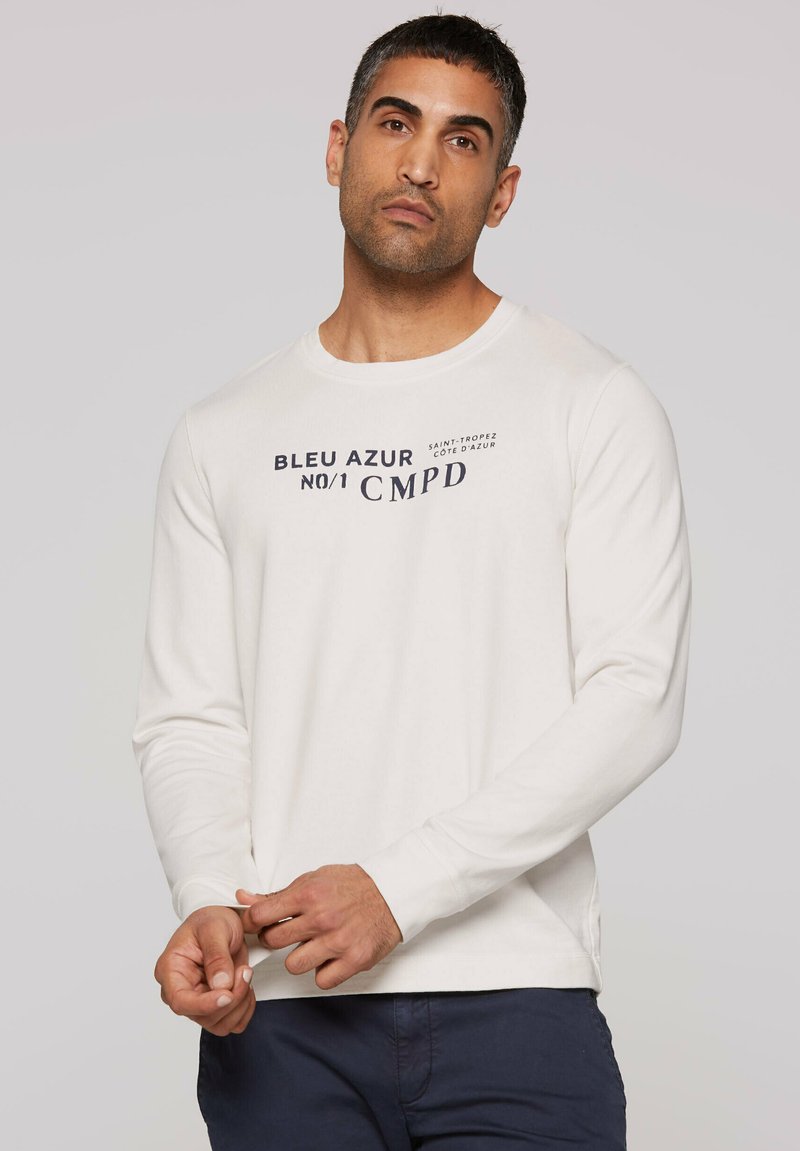 Long-sleeve, off-white t-shirt made of cotton blend, featuring navy text and a crew neck. Hands adjusting the hem.