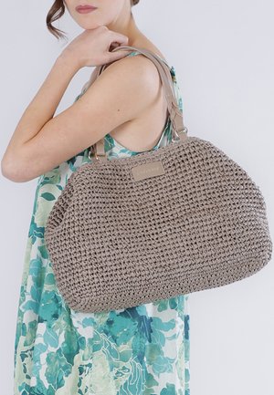 Vrouw in mouwloze bloemenjurk met grote beige gevlochten handtas over de schouder tegen een effen achtergrond.