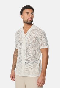 Chemise à manches courtes en dentelle blanche avec des motifs floraux complexes, fermeture à boutons sur le devant et col. Texturée et semi-transparente.