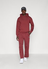 Bordeauxfarbene Kapuzenjacke und Hosen-Set mit dezentem Logo; Baumwollstoff, lockere Passform, passende Farbe, kombiniert mit weißen Schuhen und einer dunklen Mütze.