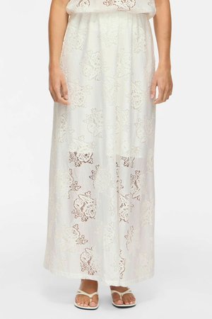 Femme portant une jupe maxi en dentelle florale blanche associée à des sandales blanches plates, se tenant devant un fond clair uni.