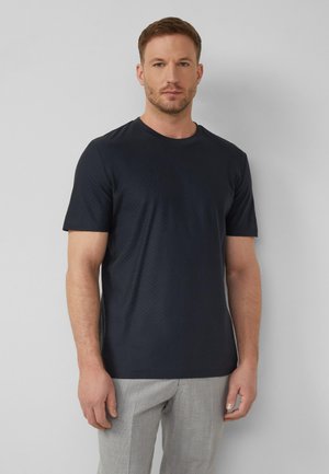 Mann trägt ein eng anliegendes schwarzes T-Shirt mit kurzen Ärmeln und hellgraue Hose, steht vor einem einfarbigen hellgrauen Hintergrund.