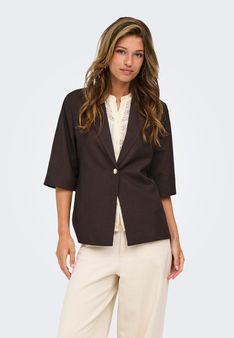 Jeune femme aux longs cheveux ondulés porte un blazer marron foncé à manches trois-quarts et un pantalon beige clair sur un fond uni.