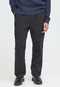 Schwarze Baumwollhosen mit gerader Silhouette, ausgestattet mit seitlichen Taschen und minimalen Nähten. Kombiniert mit schwarzen Schuhen.