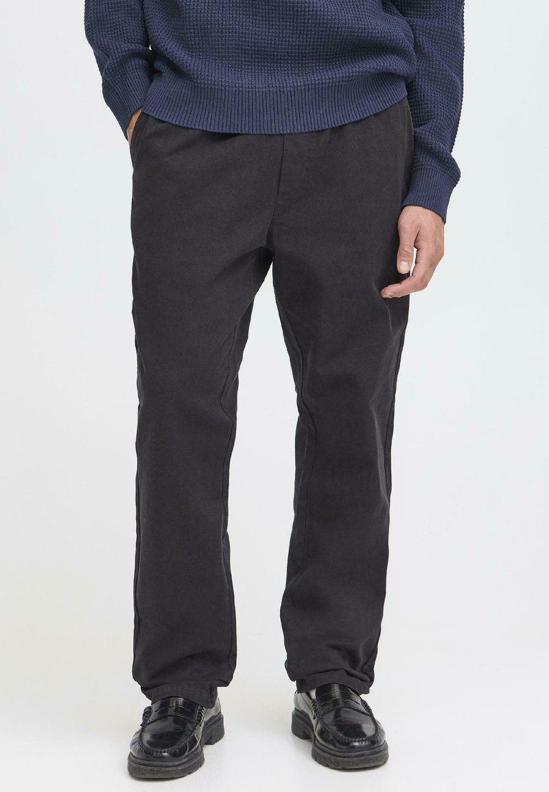 Schwarze Baumwollhosen mit gerader Silhouette, ausgestattet mit seitlichen Taschen und minimalen Nähten. Kombiniert mit schwarzen Schuhen.