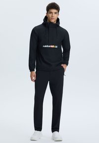 Man in een zwarte hoodie met het 'rukka' logo en vlaggen, zwarte broek en witte sneakers, staande met één hand in zijn zak.