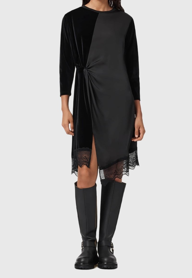Femme portant une robe noire asymétrique avec un nœud latéral froncé, un ourlet en dentelle, des manches trois-quarts et des bottes noires montant jusqu’au genou.