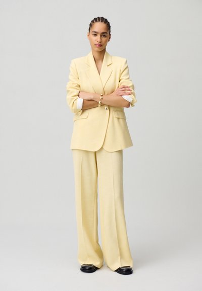 Blazer giallo su misura con ampi revers e pantaloni coordinati a gamba larga. Tessuto morbido, maniche arrotolate e scarpe nere con dettagli in argento.