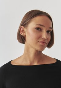 Top negru scurt cu un decolteu larg. Subiectul are o frizură bob dreaptă și poartă cercei cercuri din aur. Textura pielii este netedă, machiaj natural.