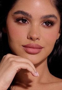 Zbliżenie twarzy z gładką, jasnobrązową cerą, pełnymi ustami w matowym odcieniu nude, ciemnymi włosami, wyrazistymi ciemnymi oczami i zadbanymi brwiami.