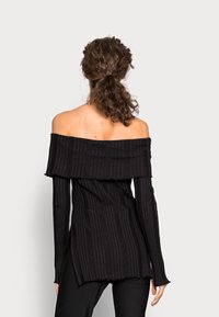 Žena s kudrnatými vlasy staženými vzadu, oblečená v černém pleteném topu s odhalenými rameny a černých kalhotách, stojící před jednobarevným světlým pozadím.